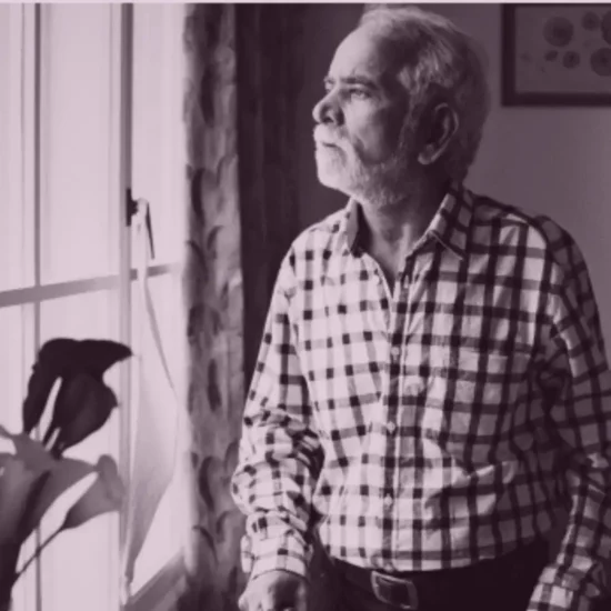 An elderly person sitting alone at home by a window, looking out quietly
