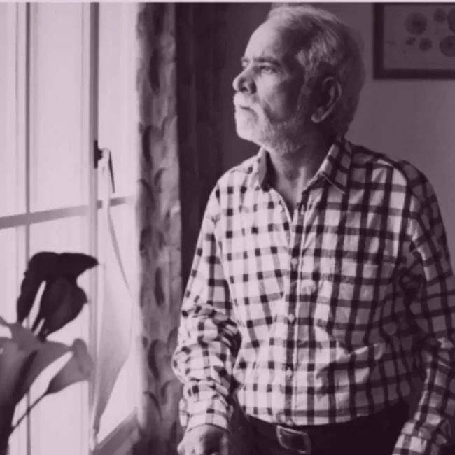 An elderly person sitting alone at home by a window, looking out quietly